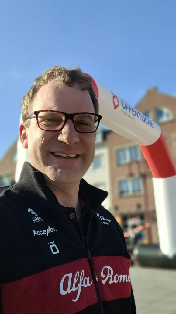 Ein Mann mit Brille und Alfa Romeo-Rennjacke lächelt für ein Selfie vor einem aufblasbaren Torbogen mit dem Schriftzug „Ostfriesische Oldenburg“, im Hintergrund sind Gebäude zu sehen.